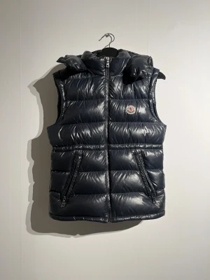 Mörkblå dunväst från Moncler - Snygg mörkblå dunväst från Moncler med glansig finish och vadderad design. Västen har huva, två framfickor med dragkedja och klassisk Moncler-logga på bröstet. Perfekt för lager-på-lager och streetstyle. Fodret har en cool serietidningsdetalj. Den passar mig som är 180cm å väger 65kg
