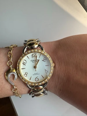 Guld- och silverfärgad Omax klocka - Snygg Omax armbandsklocka med rund urtavla, guldfärgade detaljer och glittrande stenar runt boetten. Länken är i både guld- och silverfärg med stora, blanka länkar. Urtavlan är vit med guldfärgade siffror och visare. Quartz-urverk och vattentålig. Oanvänd, endast provad