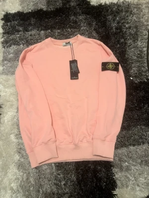 Ljusrosa sweatshirt från Stone Island - Säljer en ljusrosa sweatshirt från Stone Island med klassisk rund hals och ribbade muddar. Tröjan har den ikoniska Stone Island-patchen på vänster ärm och är tillverkad i mjuk bomull. Perfekt för dig som vill ha en clean och stilren look.