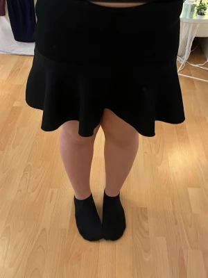 Svart volangkjol från H&M, storlek 36 - Säljer en svart kortkjol från H&M i storlek 36. Kjolen har en snygg volangkant nedtill och är tillverkad i ett strukturerat syntetmaterial. Den har en diskret dragkedja bak för enkel på- och avtagning. Perfekt för dig som gillar stilrena och trendiga plagg.