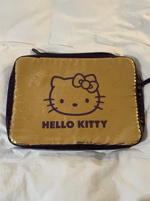 Hello Kitty datorväska  - Datorväska guldig med paljetter från Hello Kitty med lila kanter. Köpt på sellpy för 500kr. Funkar för en vanlig Apple dator. Fråga om mått så fixar jag 
