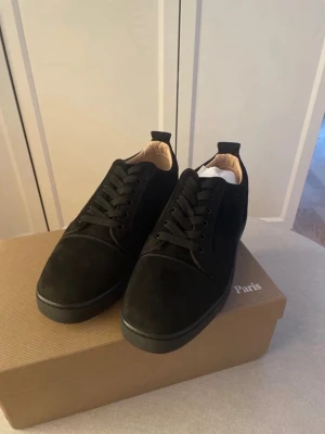 Christian Louboutin - Säljer ett par svarta sneakers från Christian Louboutin i mocka med klassiska röda sulor och diskret broderad logga på hälen. Skorna har rund tå, svart snörning och en stilren design som sticker ut med den ikoniska röda undersidan.