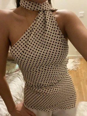 Prickig halterneck topp med scarf - Säljer en beige halterneck topp med svarta prickar och matchande scarf runt halsen. Toppen är figurnära och har en ärmlös design med asymmetrisk axel. Materialet känns mjukt och har ett lätt glansigt fall, troligen polyester eller satin.