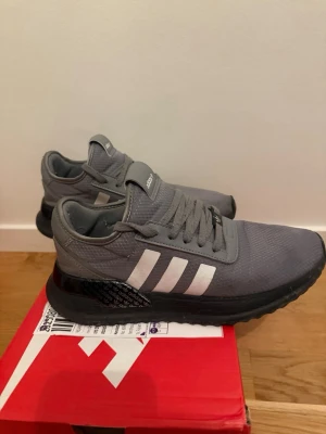 Grå adidas sneakers med vita ränder - Säljer ett par grå adidas sneakers med klassiska vita ränder på sidorna och svart sula. Skorna har snörning och är tillverkade i syntetmaterial med mesh-liknande struktur. Perfekta för dig som gillar sportig stil och vill ha bekväma skor till vardagen.