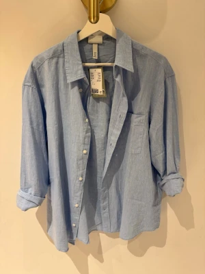 Ljusblå linneskjorta från H&M - Säljer en stilren ljusblå skjorta i linne från H&M. Skjortan har klassisk krage, knäppning framtill och en bröstficka. Ärmarna är långa och kan rullas upp för en mer avslappnad look. Perfekt för en fräsch och enkel stil.