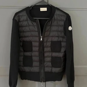 Svart cardigan  från Moncler - Säljer en svart pufferjacka från Moncler med quiltad front och fyra fickor. Jackan har långa ärmar i stickad kvalitet och klassisk Moncler-logga på ena ärmen. Dragkedja framtill och ribbade muddar för en clean look. Perfekt för dig som vill ha något stilrent och bekvämt.