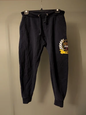 Marinblå mjukisbyxor med tryck och snörning - Marinblå mjukisbyxor med resår och snörning i midjan. På vänster ben finns ett stort tryck med text och emblem i gult, vitt och rött. Byxorna har muddar vid bensluten och är gjorda i mjukt bomullsmaterial. Perfekta för chill eller träning.