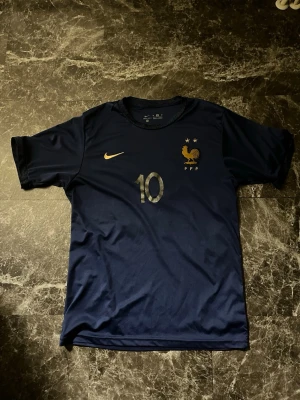 Frankrike Nike Mbappé fotbollströja - Mörkblå officiell Frankrike fotbollströja från Nike med guldiga detaljer, nummer 10 och Mbappé på ryggen. Tröjan har korta ärmar, FFF-logga och två stjärnor på bröstet. Perfekt för dig som älskar fotboll och vill stötta landslaget.
