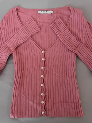 Rosa ribbad cardigan från NA-KD - Supersnygg rosa cardigan jacka från NA-KD i ribbad stickad kvalitet. V-ringad med pärlknappar framtill och långärmad passform.💞