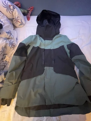 Grön och svart vindjacka från Everest - Säljer en grön och svart vindjacka från Everest med huva och dragkedja framtill. Jackan har en schysst färgblock-design och är tillverkad i slitstarkt syntetmaterial. Perfekt för dig som vill ha en snygg och funktionell jacka till höst och vinter.