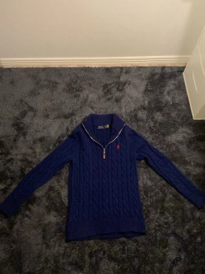 Marinblå stickad tröja Polo Ralph Lauren - Säljer en marinblå kabelstickad tröja från Polo Ralph Lauren med halv dragkedja och klassisk röd logga på bröstet. Tröjan har lång ärm, ribbade muddar och hög krage. Perfekt för kyliga dagar och snygg! OBS "Tveka inte på att skriva till mig och ge prisförslag!”