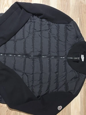 Svart  Moncler cardigan  - Säljer en svart quiltad bomberjacka från Moncler med ribbade muddar och ärmar. Jackan har dragkedja framtill och klassisk Moncler-logga på ärmen. Materialmix med polyester på kroppen och stickade detaljer på krage och ärmar. Perfekt för dig som vill ha en clean och stilren look.