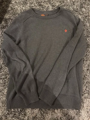 Mörkgrå stickad tröja från Morris  - Mörkgrå stickad tröja från Svea med rund halsringning och diskret röd logga broderad på bröstet. Tröjan har långa ärmar och ribbade muddar vid ärmslut och nederkant. Perfekt för lager-på-lager under kyliga dagar.