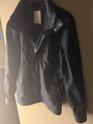 Svart vindjacka från J.Lindeberg - Snygg svart vindjacka från J.Lindeberg med dragkedja och tryckknappar. Jackan har hög krage, diskret logga på ärmen och praktiska fickor framtill. Tillverkad i lätt polyester, perfekt för blåsiga dagar. Passformen är normal och jackan är långärmad.