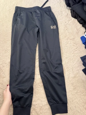 Svarta mjukisbyxor från EA7 - Säljer ett par svarta mjukisbyxor från EA7 Emporio Armani med resår i midjan och muddar vid bensluten. Byxorna har två sidofickor och en liten EA7-logga i vitt på ena benet. Perfekta för chill eller träning.