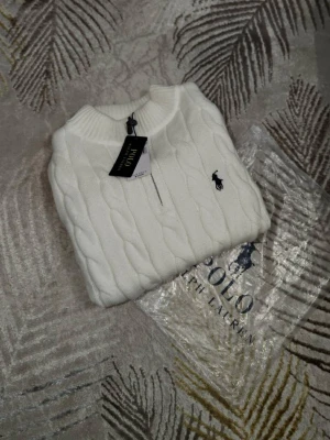 Vit kabelstickad tröja Polo Ralph Lauren - Vit kabelstickad tröja från Polo Ralph Lauren med halv dragkedja och klassisk svart broderad logga på bröstet. Tröjan har ribbade muddar och hög krage, perfekt för lager på lager under höst och vinter.