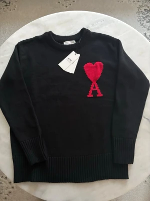 Svart stickad tröja från Ami - Svart stickad tröja från Ami med rund hals och ribbade muddar. På bröstet finns ett rött hjärta och bokstaven A, som är typiskt för Ami de Coeur. Perfekt för dig som gillar stilrena plagg med en twist. Kan diskutera pris 