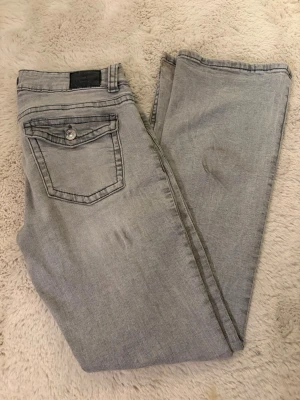 Grå jeans från Perfect Jeans - Snygga grå jeans från Gina Tricot, perfect Jeans i klassisk femficksmodell med silverfärgade knappar och detaljer. Jeansen är bootcut passform och låg midja, tillverkade i mjukt bomullsmaterial med lite stretch för extra komfort. Små fläckar på benen men går bort i tvätt, tvättas innan köp.