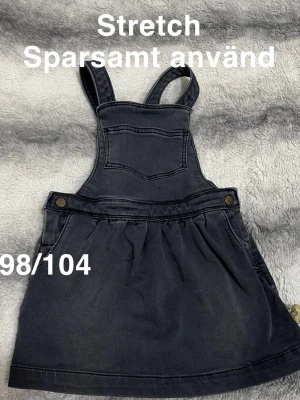 Svart jeanskjol med hängslen - Säljer en svart jeanskjol med hängslen och bröstficka. Kjolen har knappdetaljer i sidorna och är stretchig för extra komfort. Perfekt för en cool och avslappnad look.🌸 Sparsamt använd. Strechig. Storlek 98/104 