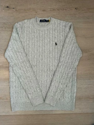 Grå kabelstickad tröja Polo Ralph Lauren - Säljer en klassisk grå kabelstickad tröja från Polo Ralph Lauren. Tröjan har rund hals, ribbade muddar och det ikoniska broderade logotypen på bröstet. Perfekt för lager på lager under höst och vinter.