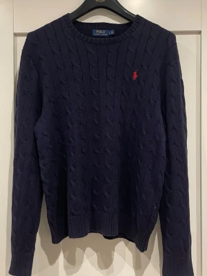 Mörkblå kabelstickad tröja Polo Ralph Lauren - Kabelstickad mörkblå tröja från Polo Ralph Lauren med klassisk röd logga broderad på bröstet. Tröjan har rund halsringning, ribbade muddar och är långärmad. Perfekt för lager-på-lager under kyliga dagar.