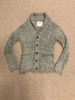 Gråmelerad kofta Abercrombie & Fitch - Säljer en gråmelerad stickad kofta från Abercrombie & Fitch. Koftan har stora svarta knappar med logga, två fickor fram och en mysig krage. Perfekt för lager på lager under kyliga dagar. Långärmad och i mjukt material. Storlek S