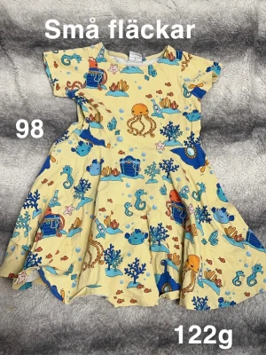 Gul klänning med havsmotiv, stl 98 - Söt gul klänning med färgglatt undervattensmönster med bläckfiskar, sjöhästar och skattkistor. Klänningen har korta ärmar, perfekt för varma dagar. Lekfull och bekväm modell som passar till sommaren.🌸 Små fläckar. Storlek 98