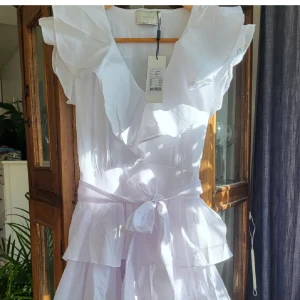 Vit volangklänning från Neo Noir - Superfin vit kortklänning från Neo Noir med volanger, omlott-v-ringning och knytband i midjan. Klänningen har flera lager och är ärmlös med breda axelvolanger. Perfekt för studenten ❤️❤️❤️ny med prislapp. Nypris 899kr