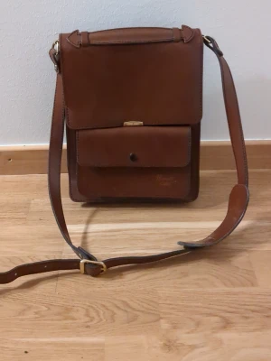 Handväska/axelremsväska  - Skinväska i portfölj-liknande stil. Bra skick förutom skador på ytan på framsidan. Går säkert att fixa för den som vill. Vet inte tillräckligt om skin för att kunna säga om det är äkta eller inte. Lagom storlek för en surfplatta. Min 13" dator är precis för stor för att få plats. 