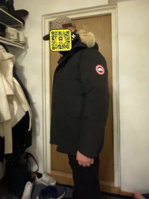 Svart dunjacka från Canada Goose - Säljer en svart dunjacka från Canada Goose med klassisk patch på ärmen och huva med snygg pälsdetalj. Jackan har knappar framtill och är perfekt för kalla dagar. Stilen är clean och passar dig som vill ha något både varmt och stilrent.