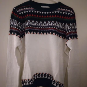 Mönstrad stickad tröja / jultröja från H&M L.O.G.G, Medium, NYSKICK - Stickad tröja / jultröja från H&M L.O.G.G. i storlek M. Tröjan är vit med flerfärgat mönster över bröst, axlar och nederkant. Rund hals och långa ärmar. Perfekt för dig som gillar klassiska vintervibbar och snygga detaljer. Nyskick, endast använd 1 gång. Felfri.