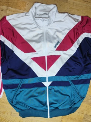 Vintage Adidas track jacket 80s/90s colorblock XL - Vintage Adidas track jacket från sent 80-tal / tidigt 90-tal. Klassisk colorblock-design i vitt, rött, marinblått och turkost. Broderad Adidas-logga på bröstet, original dragkedja och klassiska tre ränder på ärmarna. Original vintage (etiketter kvar, se bilder).                             Boxig/oversized passform – sitter snyggt även på L för oversize-look Mycket bra skick för åldern.                                     Armhåla till armhåla 69cm. Längd axel-nederkant 72cm