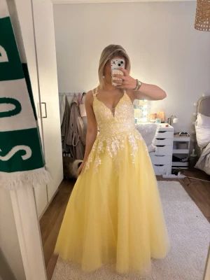 Gul balklänning - Superfin gul långklänning med v-ringning och tunna axelband som korsas i ryggen. Klänningen har vita blommiga spetsdetaljer upptill och över midjan, samt en voluminös kjol i tunt, flowy tyg. Hade den på min bal i 9an. Mina mått: midja - 69 cm höft- 82 cm bryst- 84 cm axel till höft 53 cm. Skriv om du undrar något