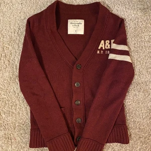 Vinröd kofta från Abercrombie & Fitch - Säljer en snygg vinröd kofta från Abercrombie & Fitch med vita ränder på ärmen och A&F-broderi på bröstet. Koftan har en v-ringad design med knappar framtill och ribbade muddar. Perfekt för en stilren look!