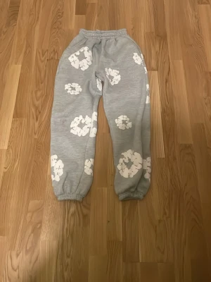 Grå mjukisbyxor från Denim Tears - Grå mjukisbyxor från Denim Tears med vita blommönster över hela byxorna. Tillverkade i mjukt bomullsmaterial, perfekta för chill dagar.