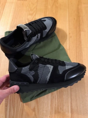 Valentino Garavani Rockrunner sneakers - Säljer ett par svarta och grå Valentino Garavani Rockrunner sneakers med camouflagemönster. Skorna har snörning, meshpaneler, detaljer i mocka och skinn samt en chunky sula med nitar på hälen. Perfekta för dig som gillar streetstyle och exklusiva märken.