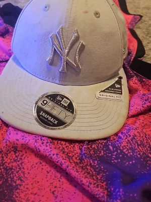 Grå New Era snapback-keps - Säljer en stilren grå snapback-keps från New Era med broderad NY-logga framtill och klassisk snapback-spänne bak. Kepsen har originalstickers kvar och är i modellen 9Fifty med slim crown och squared visor. Perfekt för dig som gillar streetwear.