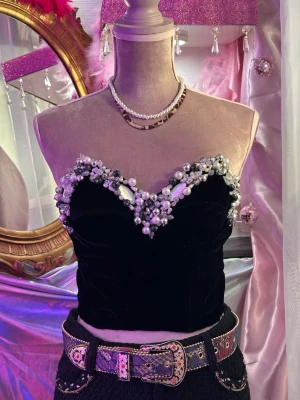 Svart sammets tubtopp med pärldetaljer - Unik svart tubtopp i sammet med hjärtformad urringning, dekorerad med pärlor och glittrande stenar längs kanten. Toppen är axelbandslös och har en lyxig känsla som verkligen sticker ut. Perfekt för dig som vill ha något extra till festen.