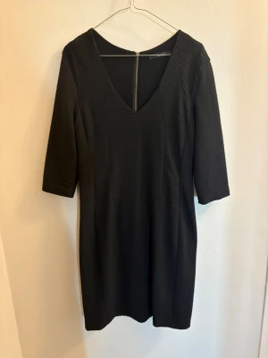 Svart ribbad kortklänning med v-ringning - Säljer en svart kortklänning med ribbad struktur och v-ringning. Klänningen har trekvartsärm och dragkedja i ryggen som detalj. Snygg och enkel modell som passar till många olika stilar.