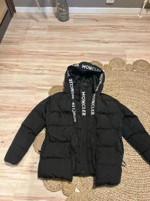 Svart Moncler pufferjacka med huva. Passar barn runt åldern 11-13 - Säljer en svart pufferjacka från Moncler med stor huva och breda snören med logga. Jackan har dragkedja, knappar och broderad Moncler-patch på ärmen. Insidan har ett unikt serietryck och klassisk Moncler-etikett. Perfekt för kalla dagar och riktigt snygg streetstil. En av Jackans dragkedja funkar inte men det är därför det sälj på ett billigt pris 