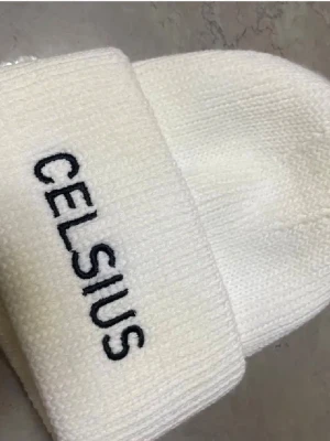 Vit stickad mössa från Celsius - Säljer en stilren vit stickad mössa från Celsius med svart broderad logga framtill. Mössan har vikt kant och är ribbstickad för extra komfort och snygg passform. Perfekt accessoar för kalla dagar.