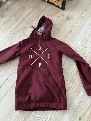 Vinröd skidjacka från Dope - Säljer en vinröd skid/snowboard jacka  från Dope med stor vit logga på bröstet. Jackan har huva med snörning, magficka och elastiska muddar. Perfekt för dig som vill ha en snygg och funktionell jacka till skidbacken. Passar både tjejer och killar.