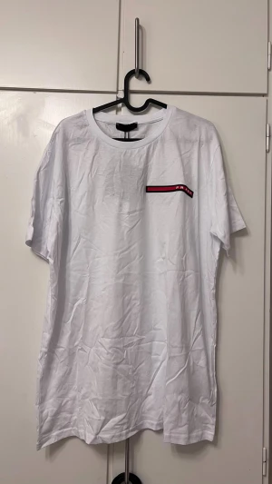 Vit t-shirt  röd detalj - Snygg vit t-shirt klassisk rund hals och korta ärmar. På bröstet finns en röd och svart detalj med logo som ger en clean men exklusiv vibe. Tillverkad i mjuk bomull för skön känsla. Perfekt till jeans eller shorts.