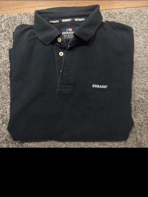 Svart pikétröja från Sebago i storlek L svartfärgad  - Klassisk svart pikétröja från Sebago med broderad logga på bröstet. Tröjan har krage, knappar framtill och är tillverkad i bomull. Långärmad modell som passar perfekt till både jeans och chinos.