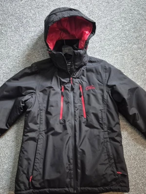 Svart skidjacka från Five season med huva - Säljer en svart skidjacka från Five season med röda detaljer och dragkedjor. Jackan har en stor huva med röd insida och flera praktiska fickor framtill. Perfekt för kalla dagar och riktigt snygg med sin kontrastfärgade design.