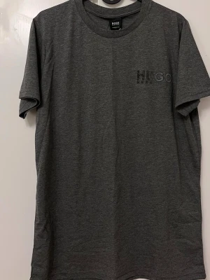 Grå t-shirt i storlek M - Snygg gråmelerad t-shirt  diskret logga på bröstet. Klassisk rund halsringning och korta ärmar. Perfekt för en clean och stilren look. Materialet är mjukt och skönt, passar till allt.