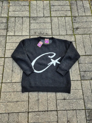 Corteiz - Mysig mörkgrå stickad tröja från PWS med stor vit C och stjärna på framsidan. Rund halsringning och ribbade muddar vid ärmslut och nederkant. Liten broderad logga på ärmen för extra detalj. Perfekt för kalla dagar.