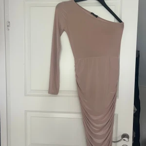Beige långklänning med oneshoulder - Säljer en beige långklänning från PrettyLittleThing med oneshoulder och lång ärm. Klänningen har draperad effekt längs kjolen och är tillverkad i ett stretchigt, mjukt material som sitter snyggt på kroppen.