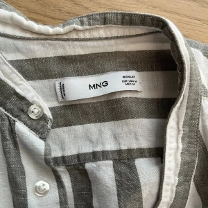 Randig linneskjorta från MNG, M - Säljer en snygg randig skjorta från MNG i storlek M. Skjortan är vit med breda grå ränder, har ståkrage och knäppning framtill. Tillverkad i luftig linneblandning, perfekt för en chill och stilren look. Långärmad och regular fit.