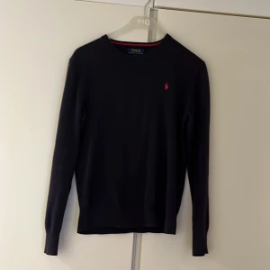 Blå Polo Ralph Lauren - Klassisk blå långärmad tröja från Polo Ralph Lauren med röd broderad logga på bröstet. Tröjan har rund halsringning med röd detalj på insidan och är tillverkad i mjukt material. Perfekt för en stilren och enkel look.